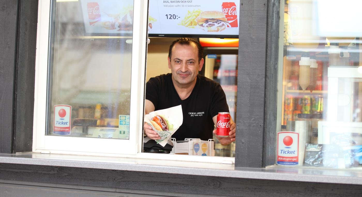 En man i en grillkiosk håller fram en hamburgare och en Coca-Cola.