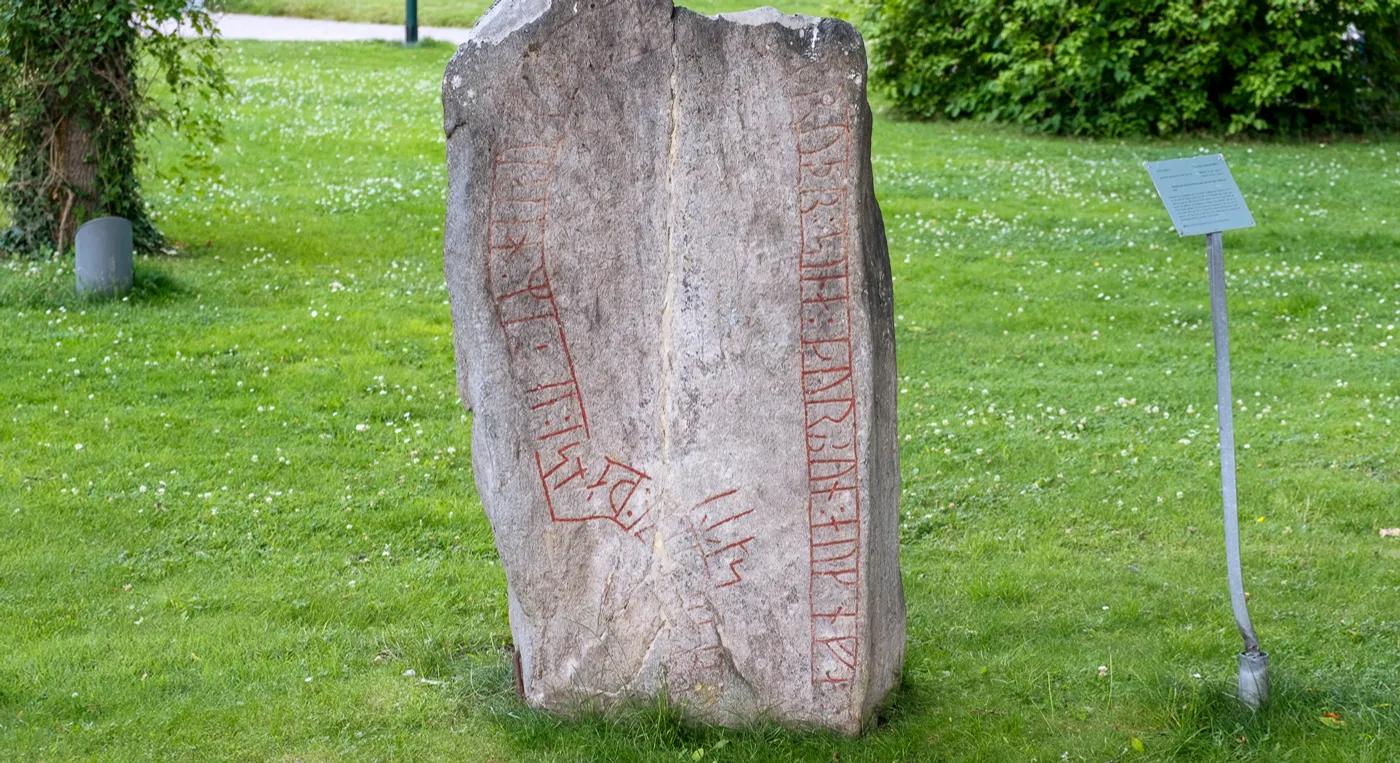 En stor runsten som ligger mitt i ett grönskande fält.