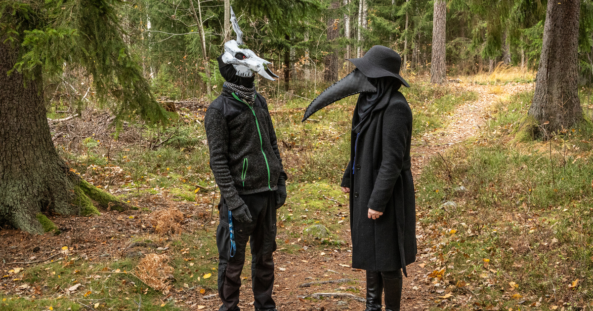 Två personer med otäcka masker står i skogen och tittar på varandra. En har ett djurkranium på huvudet och den andra en pestmask.