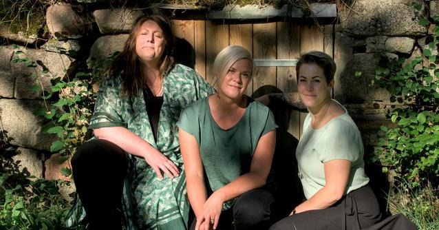 Tre kvinnor sitter  tillsammans i gräset framför en stenvägg med en trädörr