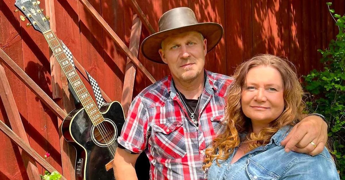 Patrik och Mikaela Rollborn i en countryinspirerad bild framför en rödmålad vägg där en gitarr är upphängd.