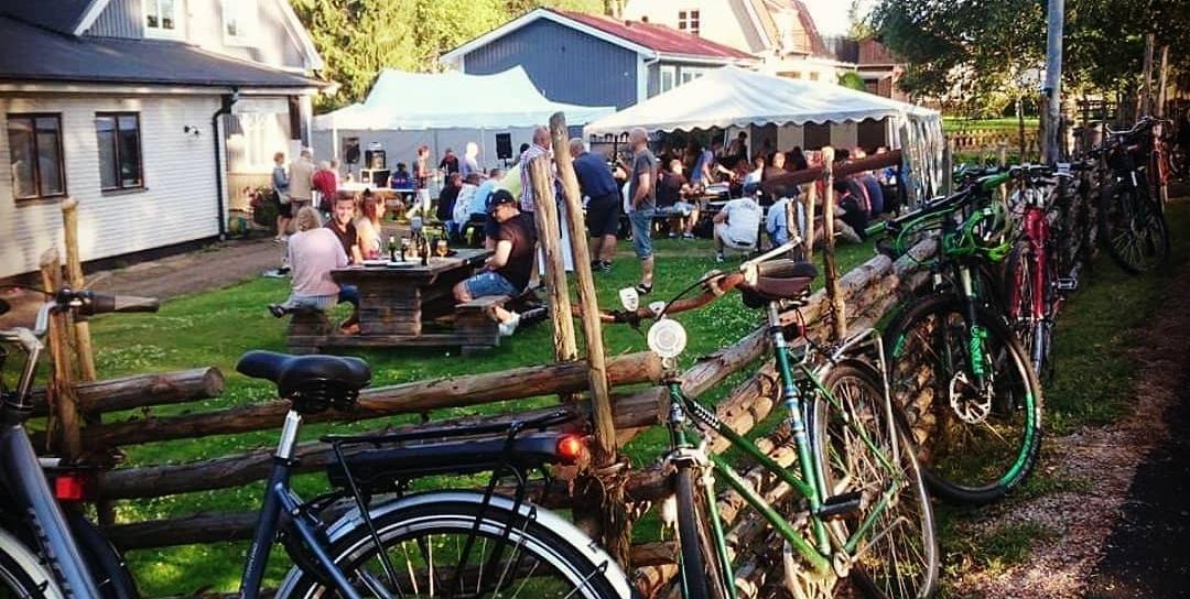 Folk sitter på bänkar i trädgården, smålandsgärdesgård där cyklar är lutade emot.