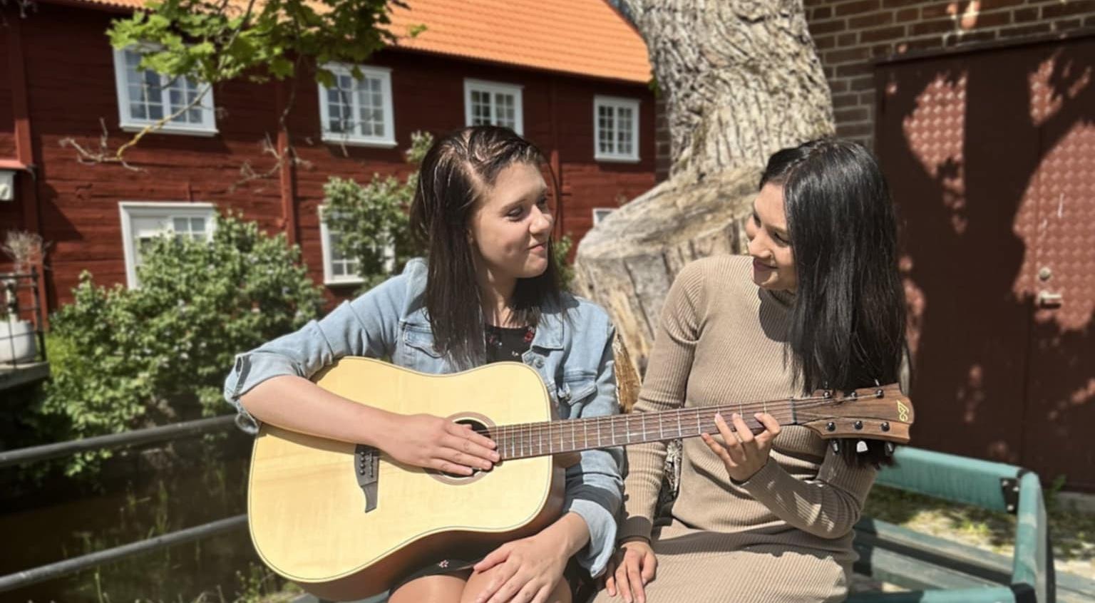 Felizia och Ramona sitterpå en bänk under ett träd och spelar på en gitarr.