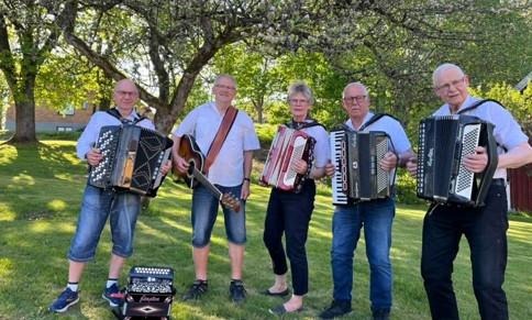 Fyra glada män och en glad kvinna med dragspel och gitarr. De står framför grönskande träd.