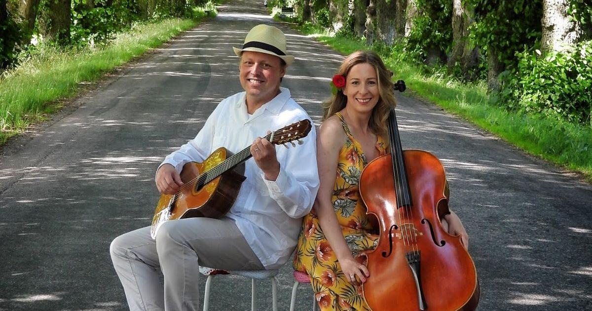 En man med en gitarr sitter på en pall på en väg, bredvid en kvinna med en cello.