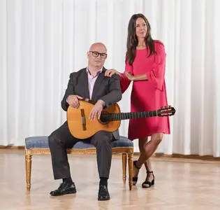 En man med gitarr sitter på en bänk, bredvid står en kvinna i klänning.